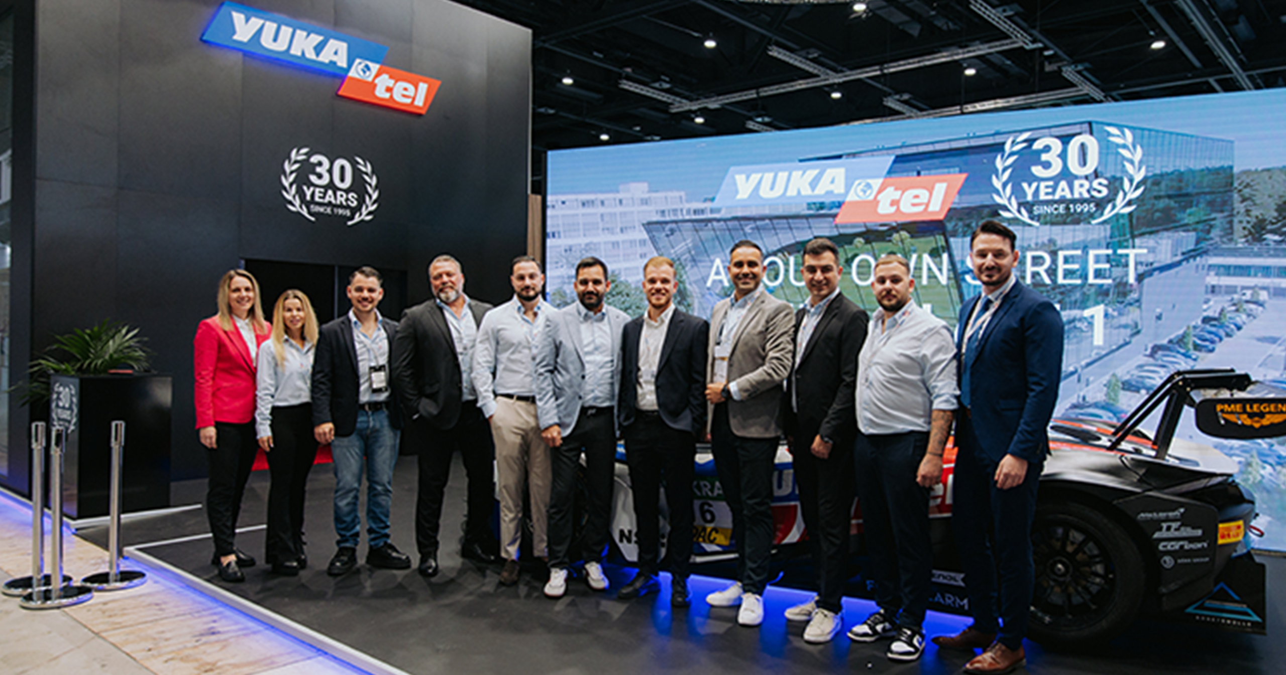 Gruppe von Menschen auf der Yukatel-Messe, 30-jähriges Jubiläum, im Hintergrund Rennwagen und Banner.