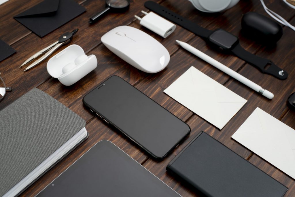 Moderne Bürogeräte auf Holzschreibtisch: Smartphone, Notizbuch, Kopfhörer, Smartwatch, Stifte, Ladegeräte und Maus.