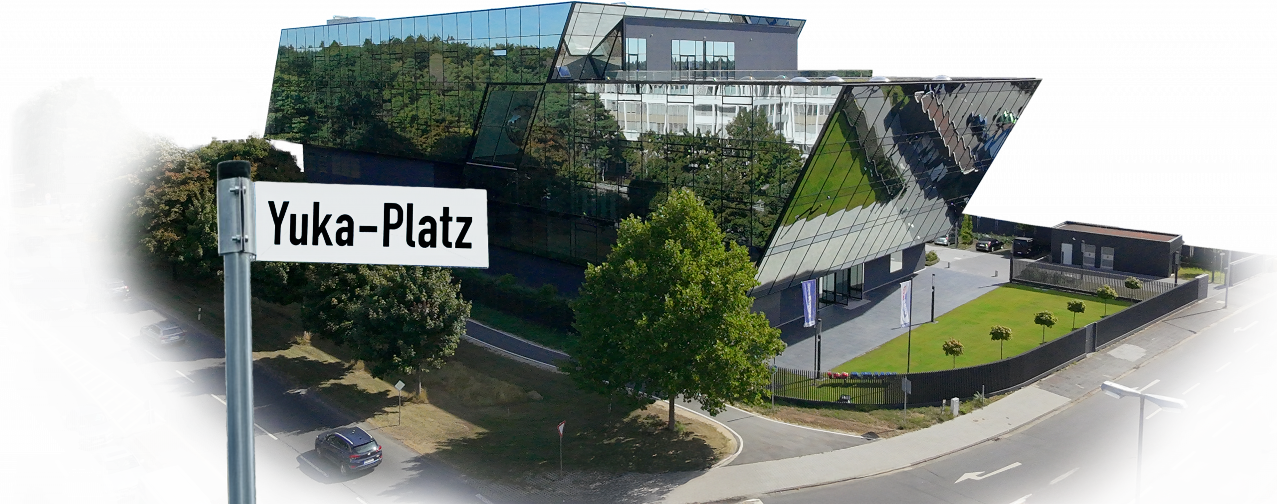 Moderne Glasfassade am Yuka-Platz mit vorbeifahrenden Autos, Straßenschild im Vordergrund.